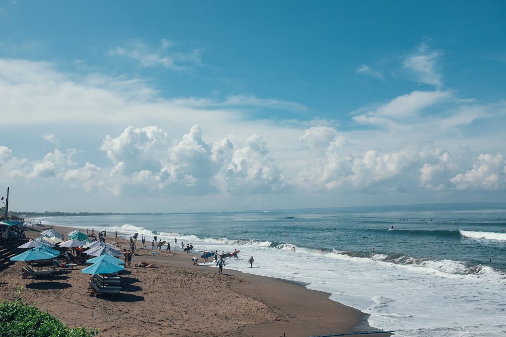 Unrecognizable tourists and athletes with surfboards on sandy beach with parasols near foamy sea under blue cloudy sky